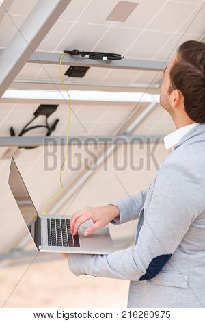 The worker of the station in the jacket checks the work of solar panels by connecting the modern laptop to the solar panel. Outdoors