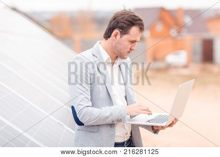 An employee of the solar station with a serious gaze holds an open modern laptop, standing next to the solar panel. Outdoors.