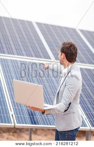 An adult man in a jacket holds an open modern laptop in his hands, and shows directional guidance on the panel of the solar battery. Outdoors .
