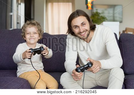 Father and son holding joysticks controllers sitting on sofa looking at camera, dad with little boy playing video games at home, man spending time with kid enjoying xbox playstation in living room