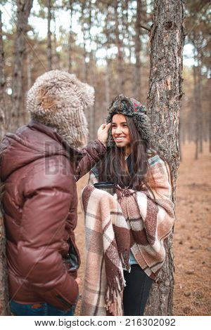 The enamored guy carefully adjusts the warm fur hat on the girl's head, the girl smiles coyly with a warm blanket. Outdoors in a cold autumn forest.