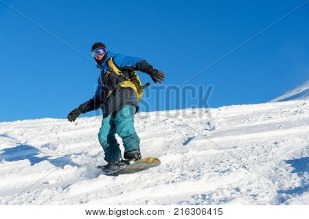 A freeboard snowboarder in a ski mask and a backpack skates over the snow-covered slope leaving behind a snow powder against the blue sky
