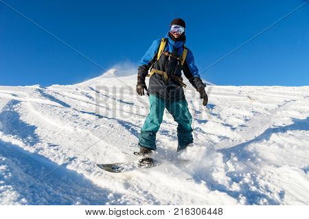 A freeboard snowboarder in a ski mask and a backpack skates over the snow-covered slope leaving behind a snow powder against the blue sky
