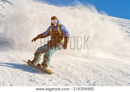 A freeboard snowboarder in a ski mask and a backpack skates over the snow-covered slope leaving behind a snow powder against the blue sky