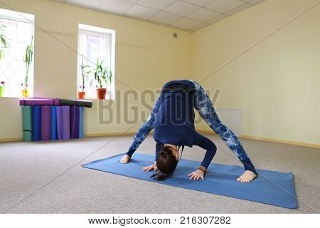 Sportswoman focused on stretch before Pilates class. Girl doing warm up for legs on mat. Female has hair pulled back, concentrated look. Athlete dressed in dark top, yoga legging trains in fitness room. Concept of online training, customizable workouts.