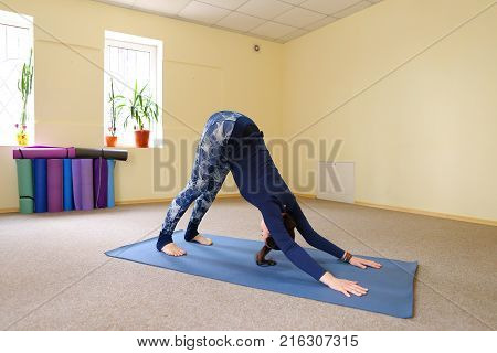 Sportswoman focused on stretch before Pilates class. Girl doing warm up for legs on mat. Female has hair pulled back, concentrated look. Athlete dressed in dark top, yoga legging trains in fitness room.  