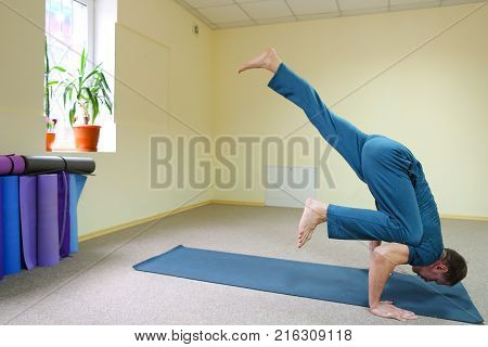 Beautiful dark-haired man performs asanas from yoga, guy sitting in room on floor on fitness mat. Inside spacious and bright, walls painted in nice yellow color, on windowsills pots with flowers. Concept of importance of keeping body in good shape, exerci