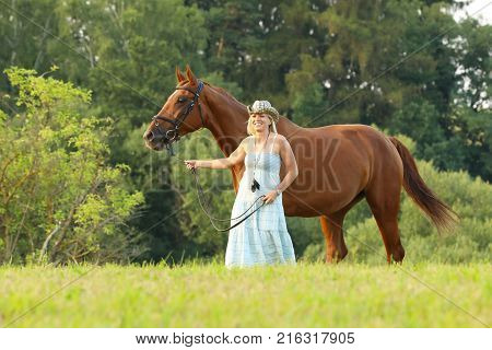 Woman in starw hat lead horse with bridle on summer afternoon