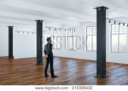 Side view of handsome young businessman standing in interior with wooden floor concrete walls columns and windows with city view. Lifestyle and workplace concept. 3D Rendering