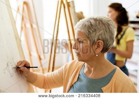 creativity, education and people concept - senior woman drawing on easel at art school studio
