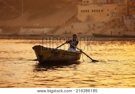 The cute Indian girl trading in ritual wreaths with flowers skillfully steering the small boat floats at sunset on golden waves of the Ganges River against the background of the ancient sacred city of Benares. India. Varanasi. February 6 2014.