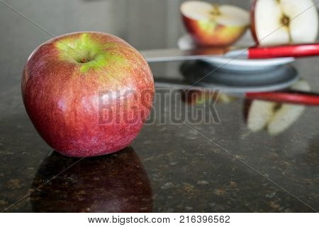 Apple in front with knife and cut apple on plate behind with reflections on granite counter.