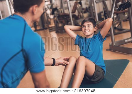 Father with his son in the same clothes in the gym. Father and son spend time together and lead a healthy lifestyle. Boy is pump the abdominals, coach helps him.