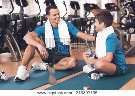 Father with his son in the same clothes in the gym. Father and son spend time together and lead a healthy lifestyle. The man and boy took a break during training. They have bath towels around necks.