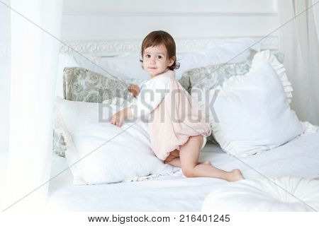 Funny and cute brunette little smiling girl playing on bed in bedroom. White interior with big bed