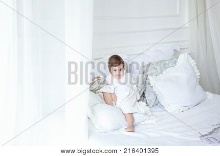 Funny and cute brunette little smiling girl playing on bed in bedroom. White interior with big bed