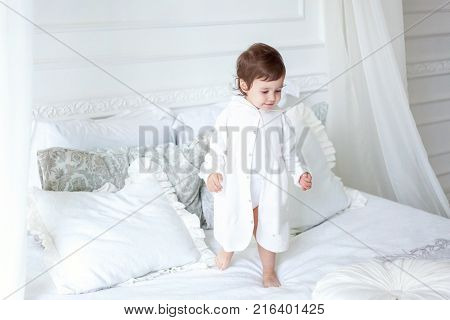 Funny and cute brunette little smiling girl playing on bed in bedroom. White interior with big bed