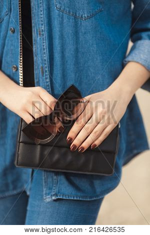 Denim outfit fashion details. Stylish woman sitting on the beach with red glitter manicure in navy jeans holding sunglasses and leather small cross body bag.