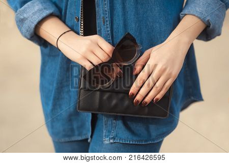 Denim outfit fashion details. Stylish woman sitting on the beach with red glitter manicure in navy jeans holding sunglasses and leather small cross body bag.