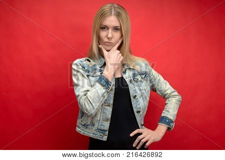 portrait on a red background in the Studio of a young long-haired woman in a denim jacket with an expressive look at the camera. expression pensive and evaluates