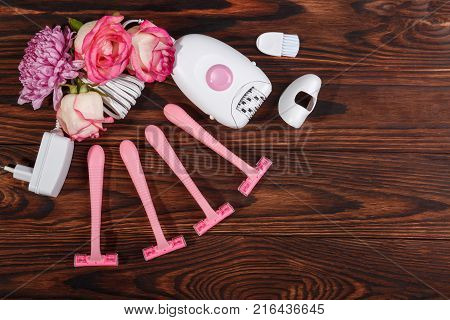 Close-up of a female epilator and four disposable pink razors with roses, lie on a wooden background, top view, free space for text.