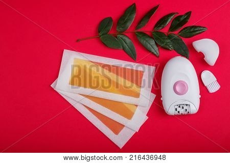 Close-up of a female epilator, wax strips, lie on a red background, next to it there is a green twig, a top view, a free place for text.