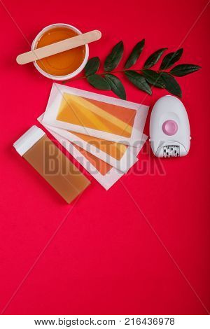 Close-up of a female epilator, wax strips, wax, wax in a cassette, lie on a red background, next to it there is a green twig, a top view, a free place for text.