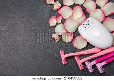 A female epilator and pink disposable razors lie near the pink rose petals, on a gray background. Hair removal, free space for text.