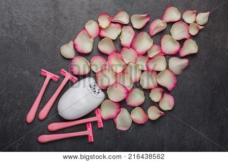 A female epilator and four pink disposable razors lie near the pink rose petals, on a gray background. Depilation.