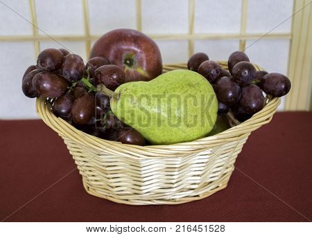 Many fruit arranged in a wicker basket on red tablecloth.