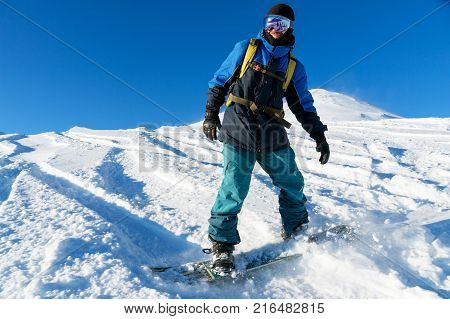 A freeboard snowboarder in a ski mask and a backpack skates over the snow-covered slope leaving behind a snow powder against the blue sky