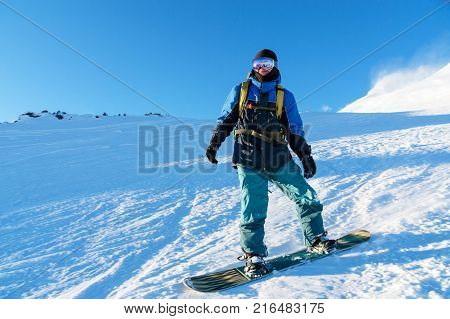 A freeboard snowboarder in a ski mask and a backpack skates over the snow-covered slope leaving behind a snow powder against the blue sky