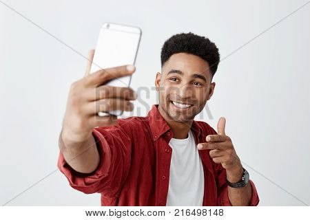Say cheese. Close up of young beautiful dark-skinned man with afro hairstyle in casual white t-shirt and red shirt smiling with teeth, holding smartphone, making selfie photo