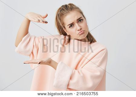 Young attractive woman wearing stylish pink long-sleeved sweatshirt showing size of something with hands while gesturing. Blonde caucasian female student posing against gray studio wall