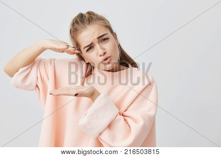 Pretty attractive charming girl wearing stylish pink long-sleeved sweatshirt showing size of something with hands, actively gesturing, frowning her face and posing against gray background.