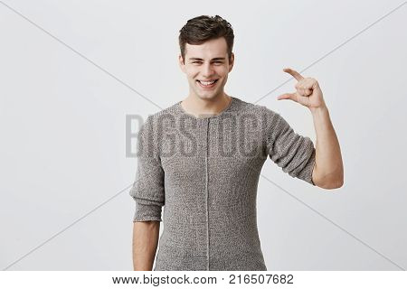 Young handsome attractive man wearing sweater showing something little with hands while gesturing and broadly smiling, demonstrating his perfect teeth. Dark-haired caucasian male model demonstrating size of something against gray studio wall
