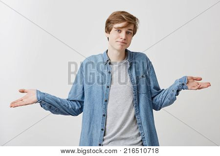Waist up portrait of confused young man with trendy hairdo and blue eyes wears demin shirt over gray t-shirt, shrugs shoulders in bewilderment as makes difficult choice or decision in his life, tries to find solution