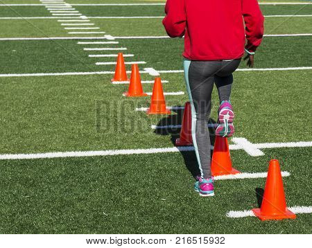A high school female athlete performs running drills over orange cones on a green turf field on a bright sunny fall afternoon wearing gray spandex.