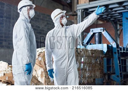 Portrait of two workers wearing biohazard suits standing in industrial warehouse of modern waste processing plant against recyclable cardboard in background