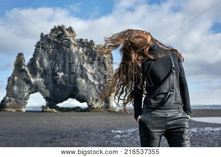 Pretty girl with windy hair stands on the volcanic beach on the background of the Hvitserkur in Iceland. She wears black jumper with dark overalls and holds hands in the pockets. Sun shine onto her.