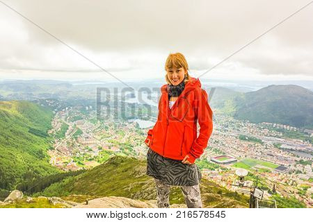 Happy young caucasian tourist in scenic norwegian landscape and popular landmark. Hiking on Mount Ulriken. Norwegian tourism highlight. Bergen urban cityscape. Traveler woman in North Europe.