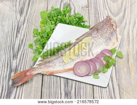 Carcass of the uncooked red cod without head with cilantro and dill twigs lemon sliced red onion on the large square white dish and bundles of the parsley on a rustic wooden surface