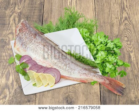 Carcass of the uncooked red cod without head sliced lemon and red onion on the large square white dish cilantro and dill on a rustic wooden surface
