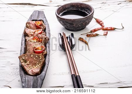 Slices of oriental spicy meat with sesame seads and pepper on long dark wooden plate with chopsticks and bowl of sauce on the white surface with cracked paint. Close up.