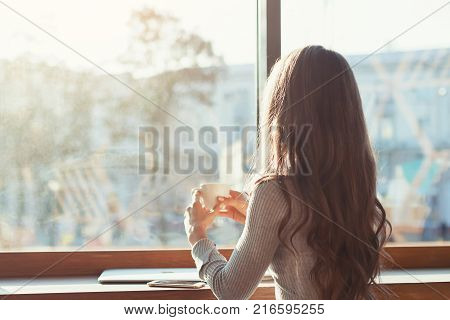 the woman in the cafe sits at a table, drinks coffee and looks at the panorama of the city outside the window. A girl in a cafe on a sunny morning