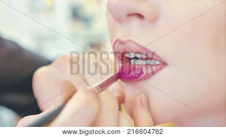 Close up of young girls's make-up process at beauty shop. Young visagiste applying pomade on model's lips. Blonde woman sitting while artist is correcting the shape of lips.