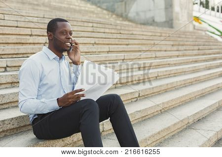 Happy black businessman talking on mobile and working with documents. African-american insuranse agent checking papers and consulting on smartphone, sitting on the stairs outdoors in urban area