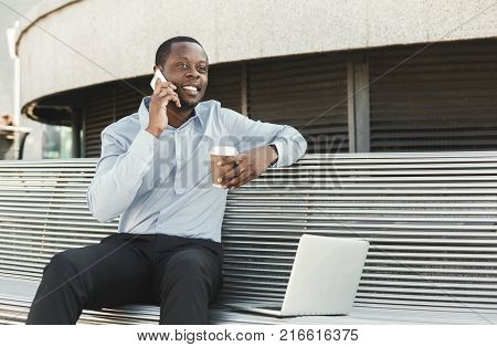 Happy black businessman working on laptop. African-american insuranse agent checking email on computer, talking on mobile, sitting on the bench outdoors near modern office center