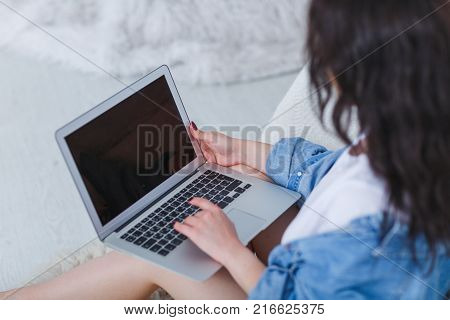 A young, beautiful bruneck woman in a denim wraps working with a laptop in her lap sitting by the bed in front of a white background