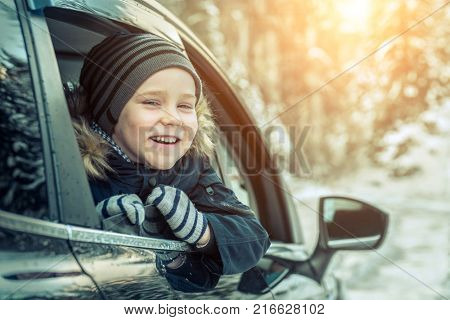 Happiness caucasian smilling boy looking out of black car window in sunny day at winter time near the forest.